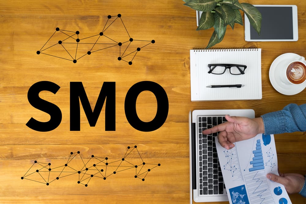 Caderno, óculos, computador e a mão de uma pessoa na direita e à esquerda, a sigla SMO, que significa Social Media Optimization.